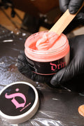 Person applying pink wax from a container labeled 'Dr' with a wooden spatula.