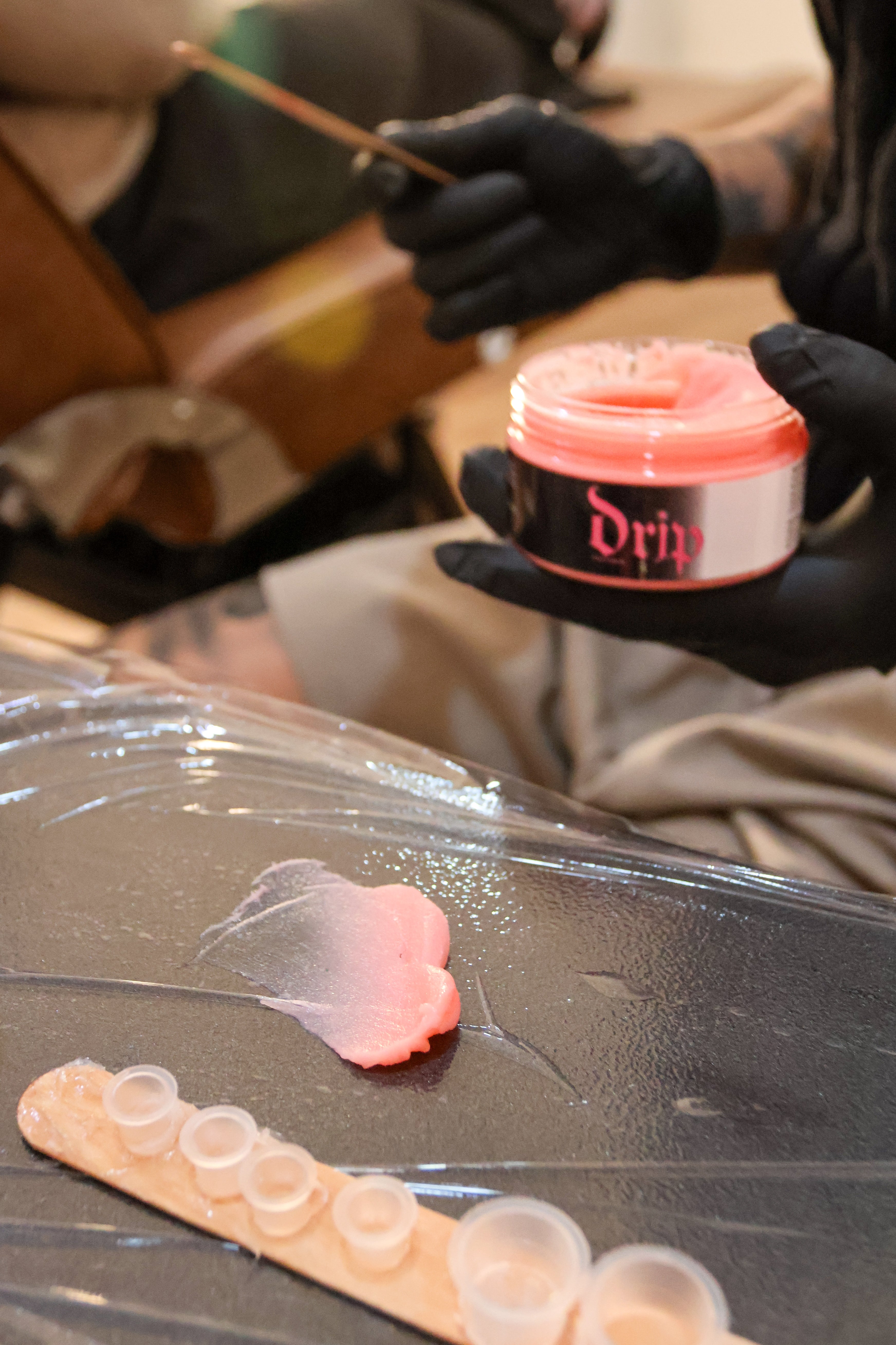 Person holding a container of pink hair dye with a tattooed arm, on a table with a clear plastic sheet.