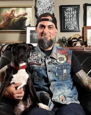 Man with tattoos and a dog sitting on a couch in a room with framed pictures and books.