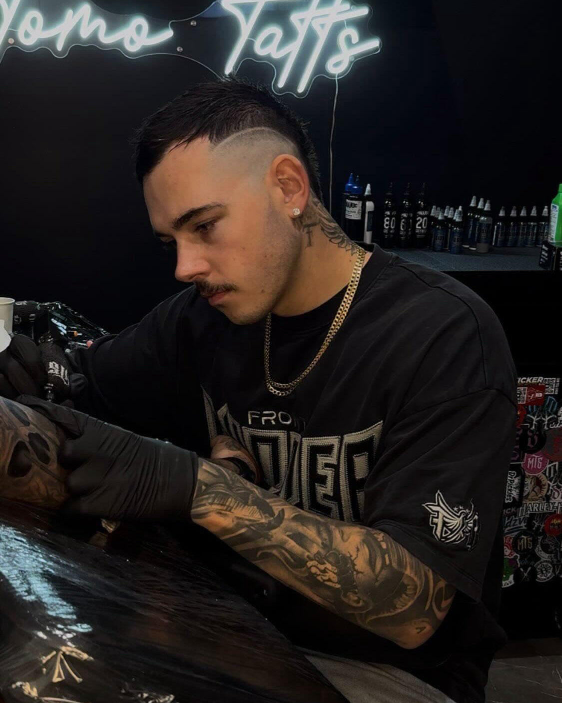 Tattoo artist working on a client in a tattoo parlor with neon signs and equipment in the background.